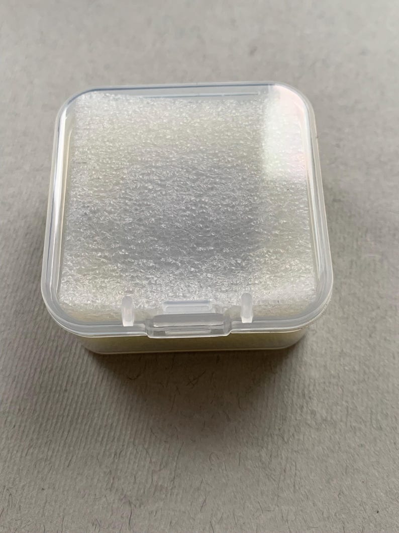 May include: A small, clear plastic box with a hinged lid. The interior is filled with a light-colored, textured foam. The box is square with rounded corners and a secure latch.