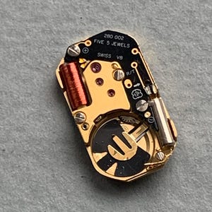 May include: A close-up of a watch movement with gold-colored components, including a coil, gears, and a black and gold rotor. The text "280 002 FIVE 5 JEWELS SWISS V8" is printed on the black circuit board.
