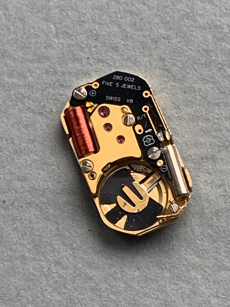 May include: A close-up of a watch movement with gold-colored components, including a coil, gears, and a black and gold rotor. The text "280 002 FIVE 5 JEWELS SWISS V8" is printed on the black circuit board.