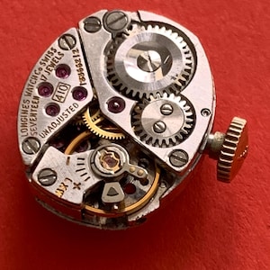 May include: Close-up of a vintage Longines watch movement with intricate gears, jewels, and the text "Longines Watch Co Swiss Seventeen 17 Jewels Unadjusted". The watch mechanism is set against a red background.