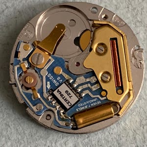 Może przedstawiać: Zbliżenie mechanizmu zegarka Certina Swiss V8. Mechanizm zegarka posiada niebieską płytkę drukowaną z logo Certina i napisem "729". Widoczne są r&oacute;wnież złote elementy metalowe i srebrna podstawa.