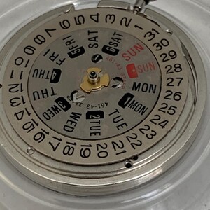May include: Close-up of a watch movement with a silver-colored dial. The dial displays the days of the week and the date, with the day of the week and date highlighted in red. A small silver crown is visible at the top.