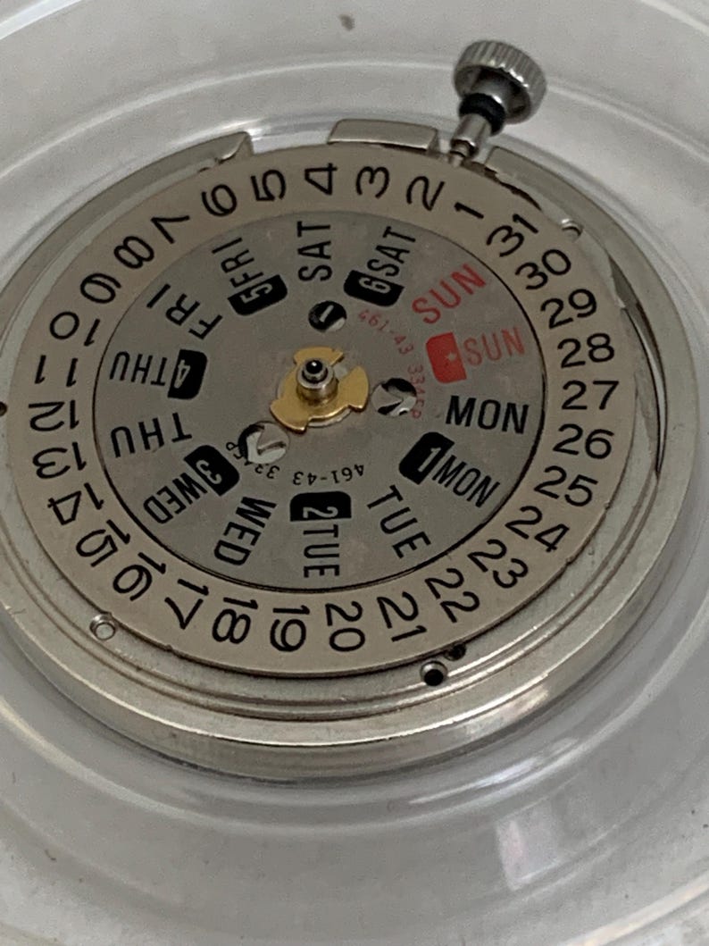 May include: Close-up of a watch movement with a silver-colored dial. The dial displays the days of the week and the date, with the day of the week and date highlighted in red. A small silver crown is visible at the top.