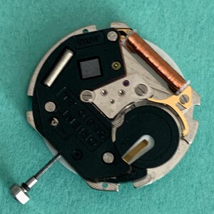May include: A detailed view of a watch movement, marked with "ORIENT 58310 21 JEWELS JAPAN". The mechanism includes a silver metal base, a black component, a copper coil, and a screw. The background is a solid teal colour.