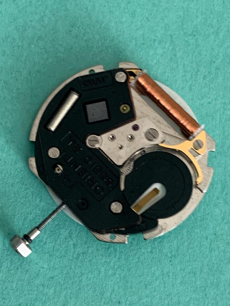 May include: A detailed view of a watch movement, marked with "ORIENT 58310 21 JEWELS JAPAN". The mechanism includes a silver metal base, a black component, a copper coil, and a screw. The background is a solid teal colour.