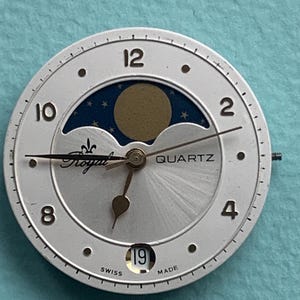 Op de afbeelding: Zilverkleurige wijzerplaat met een maanfase-ontwerp. De wijzerplaat heeft zwarte cijfers, een datumvenster met de tekst "19" en de woorden "Royal" en "Quartz". De achtergrond is licht zilver met een blauwe en gouden maanfase.