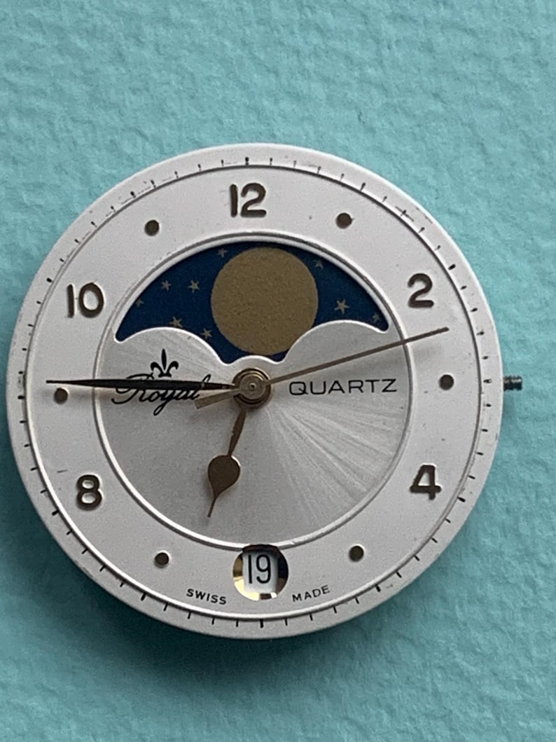 Op de afbeelding: Zilverkleurige wijzerplaat met een maanfase-ontwerp. De wijzerplaat heeft zwarte cijfers, een datumvenster met de tekst "19" en de woorden "Royal" en "Quartz". De achtergrond is licht zilver met een blauwe en gouden maanfase.