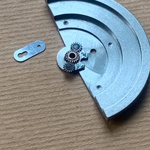 Pu&ograve; includere: Primo piano di componenti di orologio. Un pezzo di metallo argentato, semicircolare, con piccoli ingranaggi e un piccolo pezzo di metallo separato a forma di buco della serratura. Gli ingranaggi sono color rame. Lo sfondo &egrave; una superficie marrone chiaro.