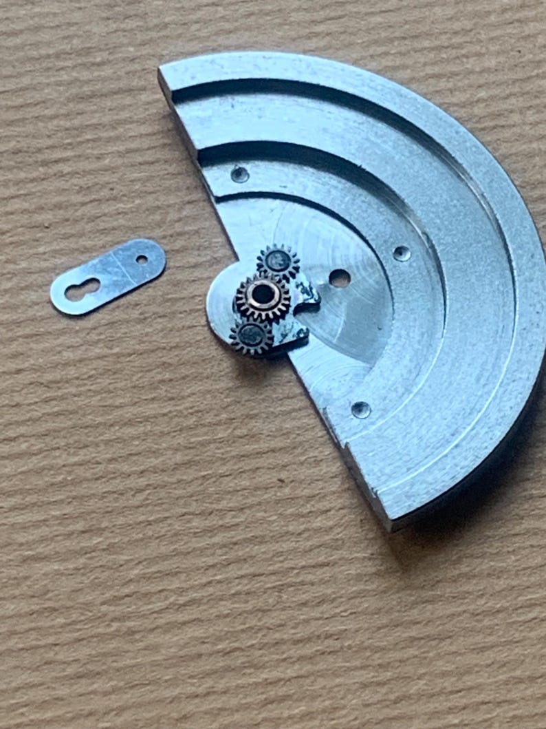 Pu&ograve; includere: Primo piano di componenti di orologio. Un pezzo di metallo argentato, semicircolare, con piccoli ingranaggi e un piccolo pezzo di metallo separato a forma di buco della serratura. Gli ingranaggi sono color rame. Lo sfondo &egrave; una superficie marrone chiaro.