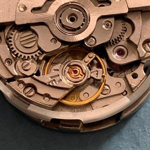 May include: Close-up of a Citizen Japan watch movement. The intricate mechanism features silver-colored gears, screws, and plates, with a gold-colored balance wheel and other components. The text "CITIZEN JAPAN JEWELS" is visible.