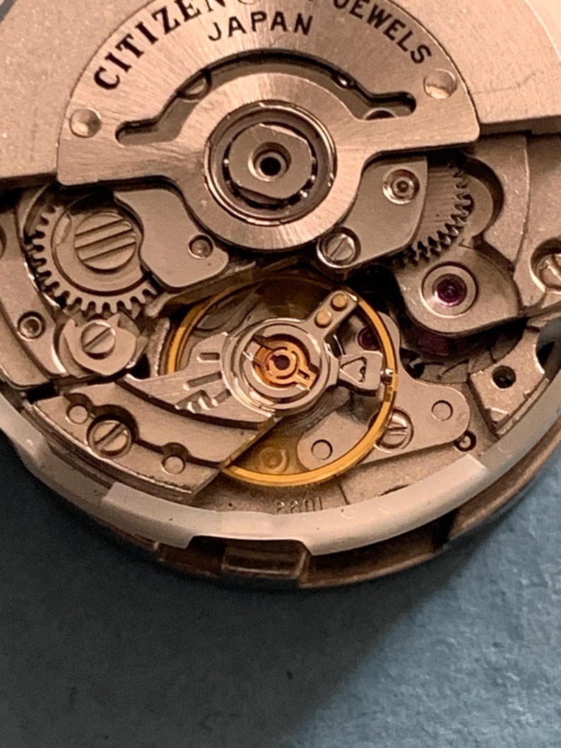 May include: Close-up of a Citizen Japan watch movement. The intricate mechanism features silver-colored gears, screws, and plates, with a gold-colored balance wheel and other components. The text "CITIZEN JAPAN JEWELS" is visible.