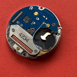 May include: A close-up of a watch movement with a blue circuit board and silver metal components. The movement has the text "SEVEN 7 JEWELS" and "UNADJUSTED". The number "4234" is visible on a metal component.