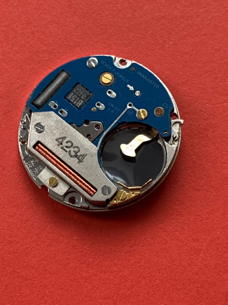 May include: A close-up of a watch movement with a blue circuit board and silver metal components. The movement has the text "SEVEN 7 JEWELS" and "UNADJUSTED". The number "4234" is visible on a metal component.