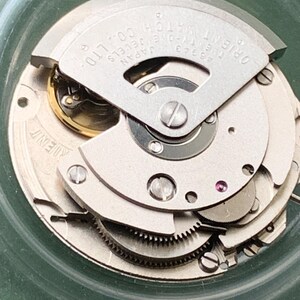 May include: A detailed view of an Orient watch movement, showcasing its internal mechanics. The silver-coloured components include gears, screws, and a jewel. The text "ORIENT WATCH CO. LTD." and "46943 JAPAN" are printed on the metal. The movement is housed in a clear, circular case.