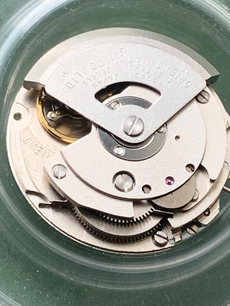 May include: A detailed view of an Orient watch movement, showcasing its internal mechanics. The silver-coloured components include gears, screws, and a jewel. The text "ORIENT WATCH CO. LTD." and "46943 JAPAN" are printed on the metal. The movement is housed in a clear, circular case.