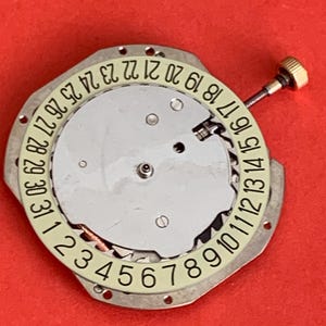 K&ouml;nnte beinhalten: Ein Uhrwerk mit silberfarbenem Zifferblatt und einem cremefarbenen Ring, der die Tage des Monats von 1 bis 31 anzeigt. Eine kleine goldfarbene Schraube ist angebracht. Der Hintergrund ist einfarbig rot.