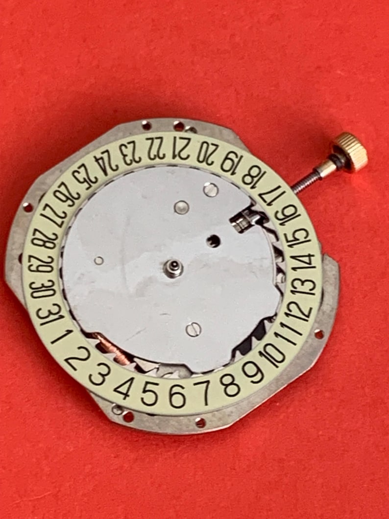 K&ouml;nnte beinhalten: Ein Uhrwerk mit silberfarbenem Zifferblatt und einem cremefarbenen Ring, der die Tage des Monats von 1 bis 31 anzeigt. Eine kleine goldfarbene Schraube ist angebracht. Der Hintergrund ist einfarbig rot.