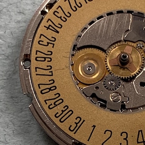 May include: Close-up of a watch movement with a gold-colored date wheel and silver-colored gears. The date wheel displays numbers from 18 to 31 and 1 to 4. The intricate mechanical components are visible.