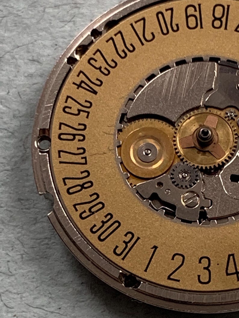 May include: Close-up of a watch movement with a gold-colored date wheel and silver-colored gears. The date wheel displays numbers from 18 to 31 and 1 to 4. The intricate mechanical components are visible.