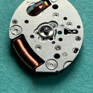 May include: Close-up of a watch movement with intricate gears, screws, and a copper coil. The silver-colored mechanism has various holes and a small, colorful component. The overall design is oval-shaped.