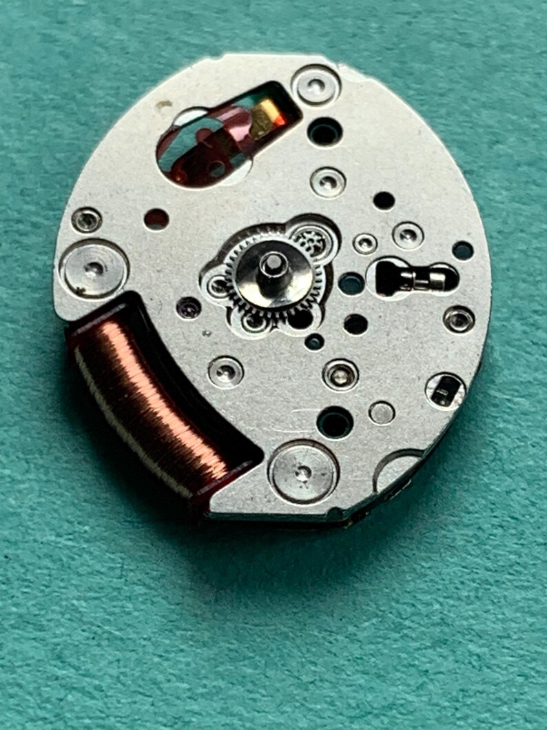 May include: Close-up of a watch movement with intricate gears, screws, and a copper coil. The silver-colored mechanism has various holes and a small, colorful component. The overall design is oval-shaped.