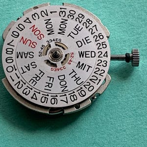 May include: A silver watch calendar dial with day and date indicators. The dial features the days of the week in English and numbers 1-31. A small crown is visible on the right side. The model number 334EG is printed on the dial.