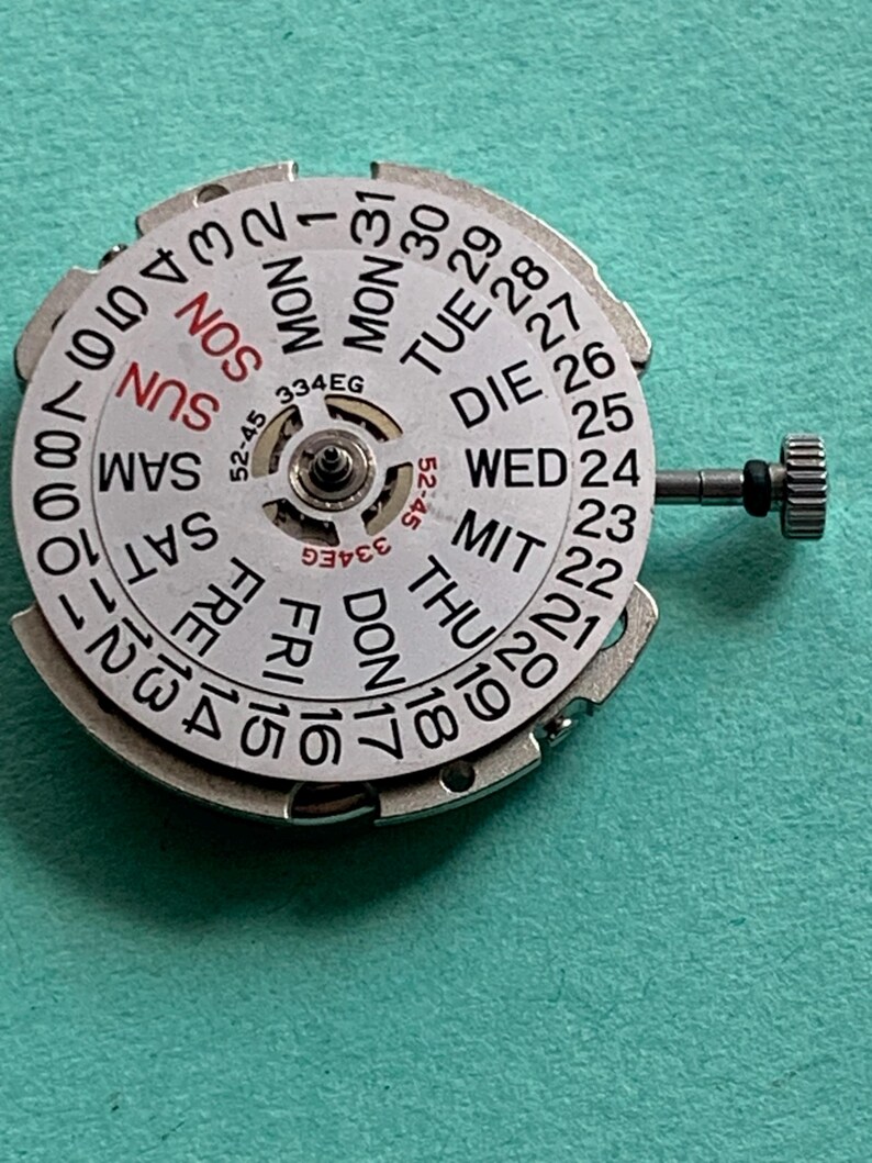 May include: A silver watch calendar dial with day and date indicators. The dial features the days of the week in English and numbers 1-31. A small crown is visible on the right side. The model number 334EG is printed on the dial.