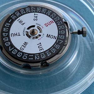May include: A watch movement with a white dial featuring black numerals and day indicators. The days of the week are printed in a circular pattern. The watch movement has a silver-coloured metal casing and a small crown.
