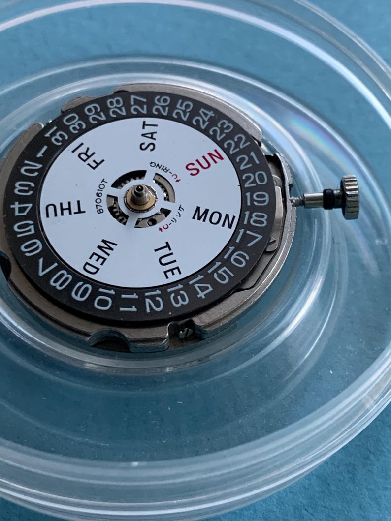 May include: A watch movement with a white dial featuring black numerals and day indicators. The days of the week are printed in a circular pattern. The watch movement has a silver-coloured metal casing and a small crown.