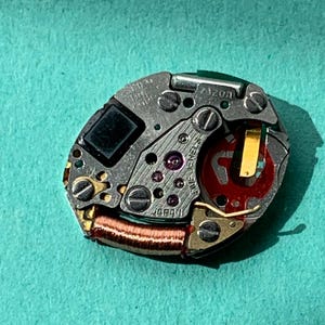 May include: Close-up of a watch movement with intricate gears, screws, and a red and gold component. The movement is marked "JAPAN" and "FIVE JEWELS". The model number "7120A" is also visible. The background is a teal color.