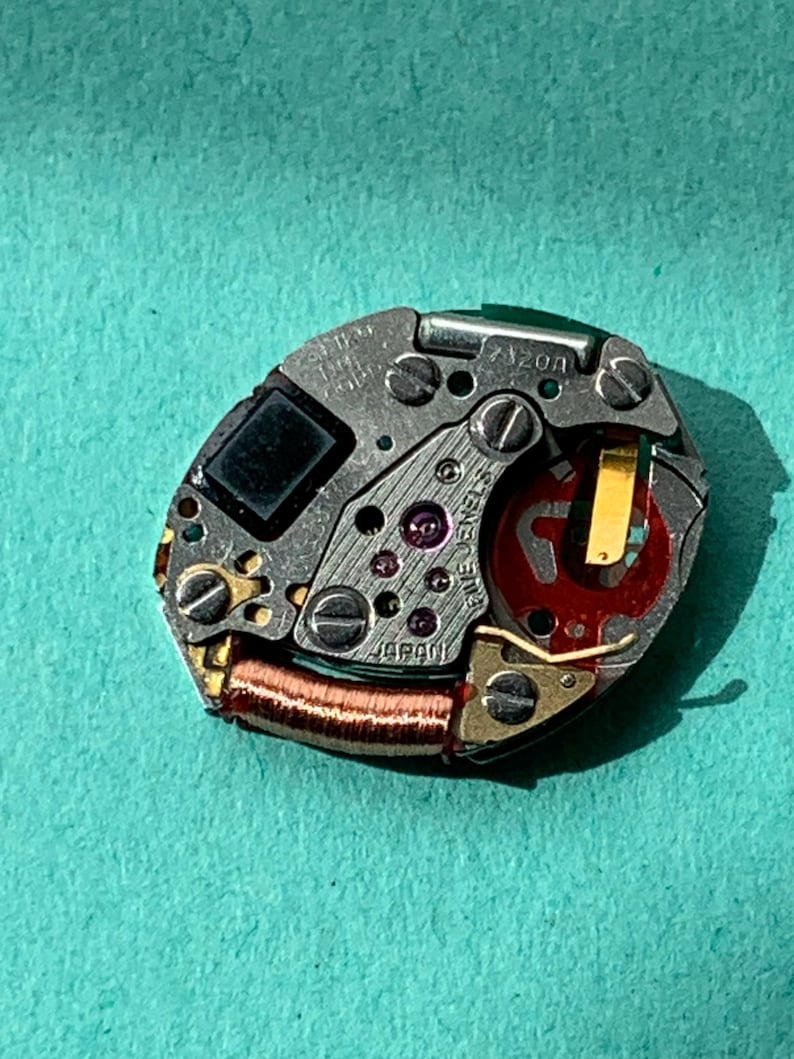 May include: Close-up of a watch movement with intricate gears, screws, and a red and gold component. The movement is marked "JAPAN" and "FIVE JEWELS". The model number "7120A" is also visible. The background is a teal color.