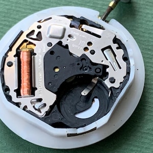 K&ouml;nnte beinhalten: Nahaufnahme eines Uhrwerks mit komplizierten mechanischen Komponenten. Der Uhrenmechanismus hat eine wei&szlig;e Basis, Metallzahnr&auml;der und eine Kupferspule. Der Text "SHIO JIRI L.D. 10 JEWELS UNADJUSTED VP32" ist sichtbar.