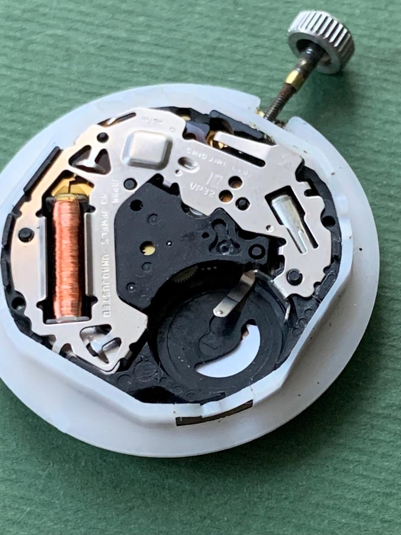 K&ouml;nnte beinhalten: Nahaufnahme eines Uhrwerks mit komplizierten mechanischen Komponenten. Der Uhrenmechanismus hat eine wei&szlig;e Basis, Metallzahnr&auml;der und eine Kupferspule. Der Text "SHIO JIRI L.D. 10 JEWELS UNADJUSTED VP32" ist sichtbar.