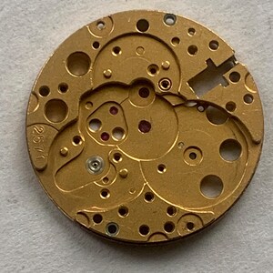 May include: Close-up of a gold-colored watch movement with intricate gears, holes, and red jewels. The number "2571" is visible on the side. This detailed mechanical component showcases the inner workings of a timepiece.