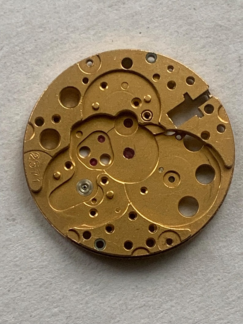May include: Close-up of a gold-colored watch movement with intricate gears, holes, and red jewels. The number "2571" is visible on the side. This detailed mechanical component showcases the inner workings of a timepiece.