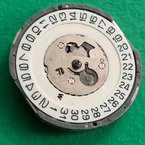 May include: A watch movement component with a white dial featuring black numerals from 1 to 31, indicating the days of the month. The component has a gray outer ring and a central mechanism with intricate metal parts.