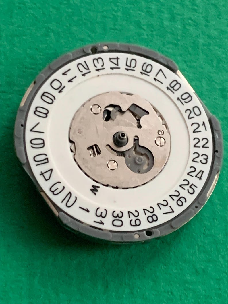 May include: A watch movement component with a white dial featuring black numerals from 1 to 31, indicating the days of the month. The component has a gray outer ring and a central mechanism with intricate metal parts.