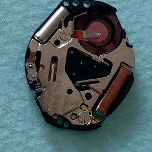 May include: A detailed view of a watch movement, showcasing intricate internal components. The mechanism includes silver metal elements, a red coil, and a black section with text. The background is a light blue surface.