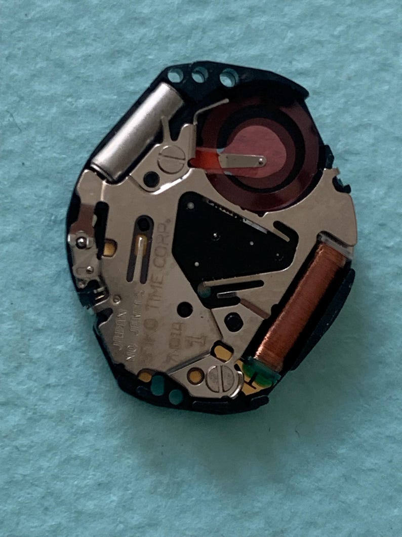 May include: A detailed view of a watch movement, showcasing intricate internal components. The mechanism includes silver metal elements, a red coil, and a black section with text. The background is a light blue surface.