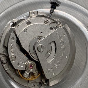 Pu&ograve; includere: Primo piano di un movimento di orologio con intricati componenti metallici. Il meccanismo presenta la scritta "ORIENT 21 JEWELS" e "JAPAN", insieme ad altri segni. &Egrave; visibile una piccola corona strutturata.
