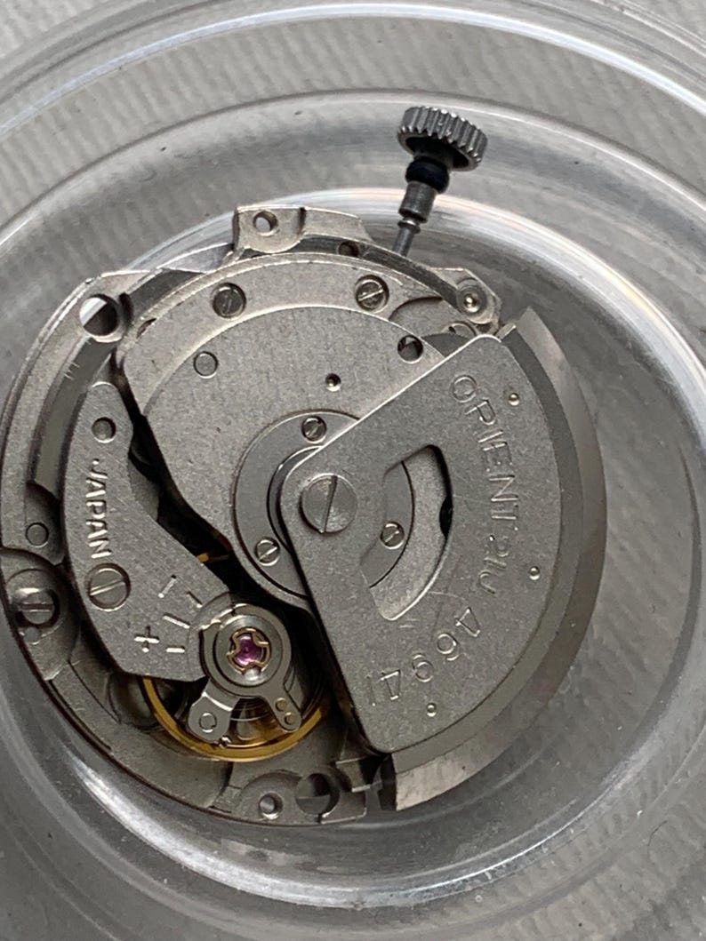 Pu&ograve; includere: Primo piano di un movimento di orologio con intricati componenti metallici. Il meccanismo presenta la scritta "ORIENT 21 JEWELS" e "JAPAN", insieme ad altri segni. &Egrave; visibile una piccola corona strutturata.