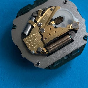 May include: A detailed view of a watch movement, featuring gold-coloured gears, a silver plate, and a green plastic frame. The text "2113A" is visible, along with other text indicating it is a 1-jewel movement made in Japan.