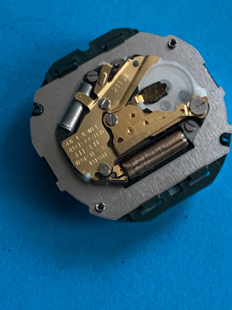 May include: A detailed view of a watch movement, featuring gold-coloured gears, a silver plate, and a green plastic frame. The text "2113A" is visible, along with other text indicating it is a 1-jewel movement made in Japan.