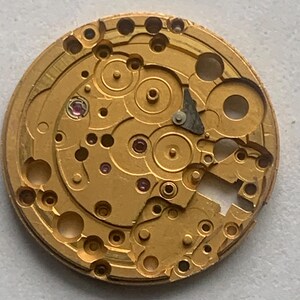 May include: Close-up of a gold-colored watch movement with intricate gears, holes, and small ruby jewels. The mechanical components are arranged in a circular pattern, showcasing the precision of the watch's internal workings.