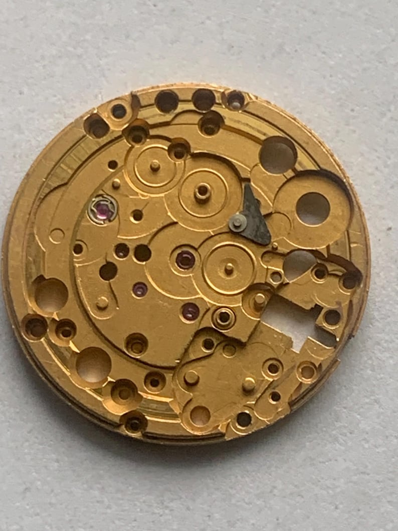 May include: Close-up of a gold-colored watch movement with intricate gears, holes, and small ruby jewels. The mechanical components are arranged in a circular pattern, showcasing the precision of the watch's internal workings.