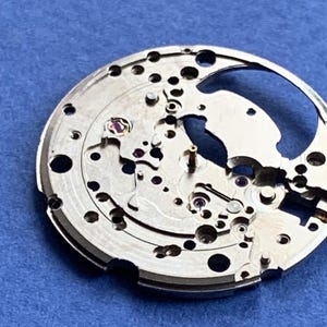 Puede incluir: Primer plano de un movimiento de reloj plateado con intrincados engranajes, tornillos y joyas. Los componentes mec&aacute;nicos est&aacute;n dispuestos en un patr&oacute;n circular, con una gran abertura. El fondo es azul liso.