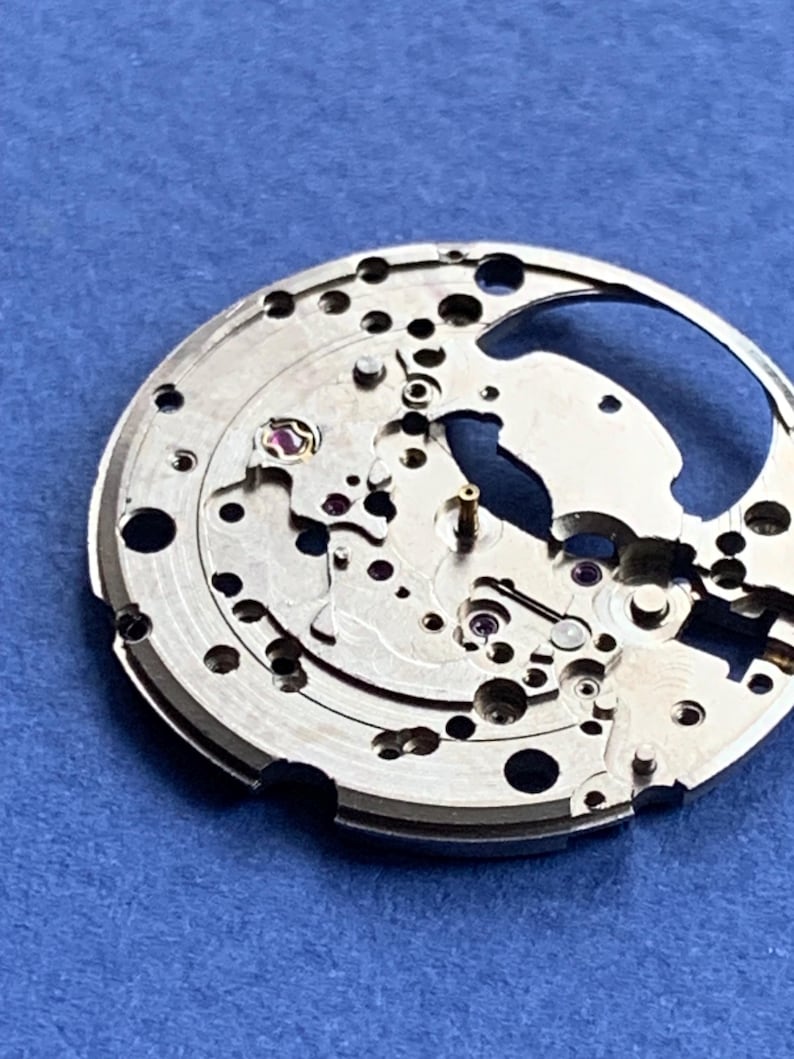Puede incluir: Primer plano de un movimiento de reloj plateado con intrincados engranajes, tornillos y joyas. Los componentes mec&aacute;nicos est&aacute;n dispuestos en un patr&oacute;n circular, con una gran abertura. El fondo es azul liso.