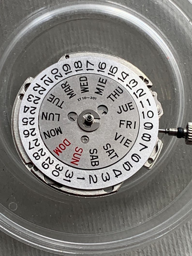 Pu&ograve; includere: Un movimento di orologio argentato con quadrante giorno e data. Il quadrante mostra i giorni della settimana in inglese e spagnolo, insieme ai numeri da 1 a 31. &Egrave; visibile anche il testo "ST 10-301".