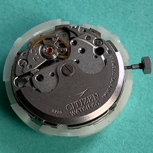 May include: A Citizen Watch Co. watch movement with the text "Twenty-One 21 Jewels" and "8200" visible. The watch mechanism is silver and gold, with a white plastic casing and a silver crown.