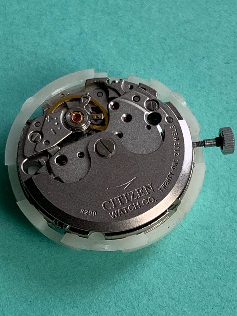 May include: A Citizen Watch Co. watch movement with the text "Twenty-One 21 Jewels" and "8200" visible. The watch mechanism is silver and gold, with a white plastic casing and a silver crown.